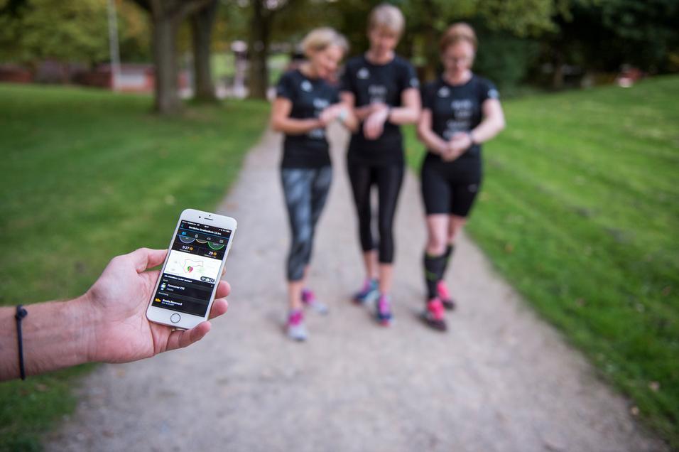 Ein Mann hält ein Smartphone mit Fitness-App, während drei Frauen in Sportkleidung auf einem Spazierweg stehen.