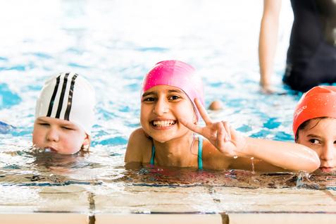 Lächelndes Mädchen mit pinker Badehaube zeigt V-Zeichen im Wasser; zwei weitere Kinder sind im Hintergrund zu sehen.